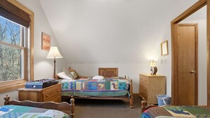 Et soveværelse med to senge, et vindue med skodder, en lampe, en trækommode og en dør.