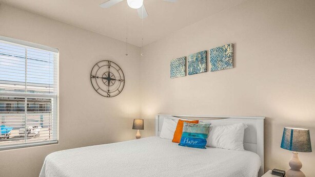 Een slaapkamer met een bed, twee nachtkastjes, een raam met zonwering, een plafondventilator en wanddecoratie.
