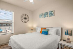 Ein Schlafzimmer mit einem Bett, zwei Nachttischen, einem Fenster mit Jalousien, einem Deckenventilator und Wandbildern.