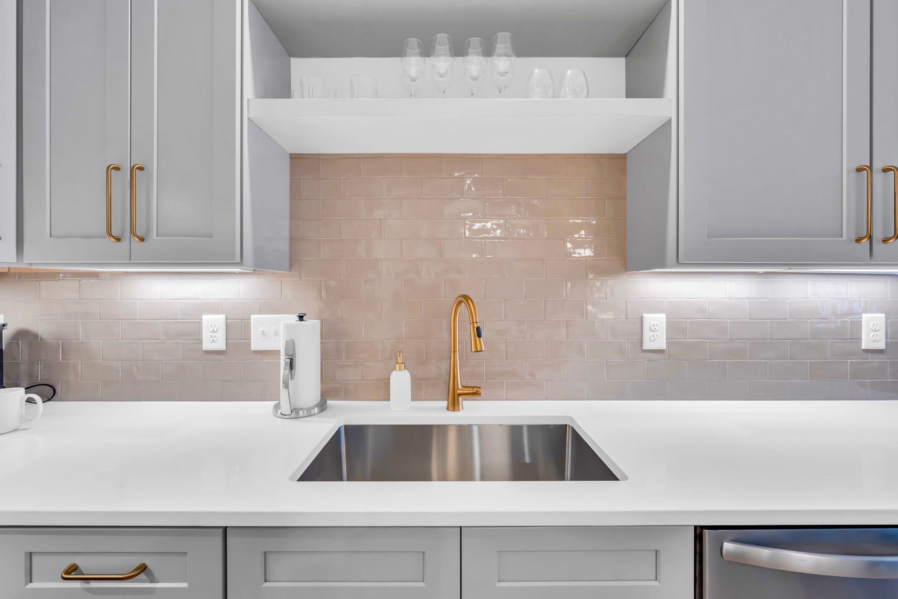 A kitchen with grey cabinets, a white countertop, a stainless steel sink, and a beige backsplash.