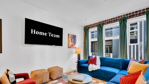 Ein Wohnzimmer mit einem blauen Sofa, roten Sesseln, einem Couchtisch und einem Fernseher, der 'Home Team' zeigt.