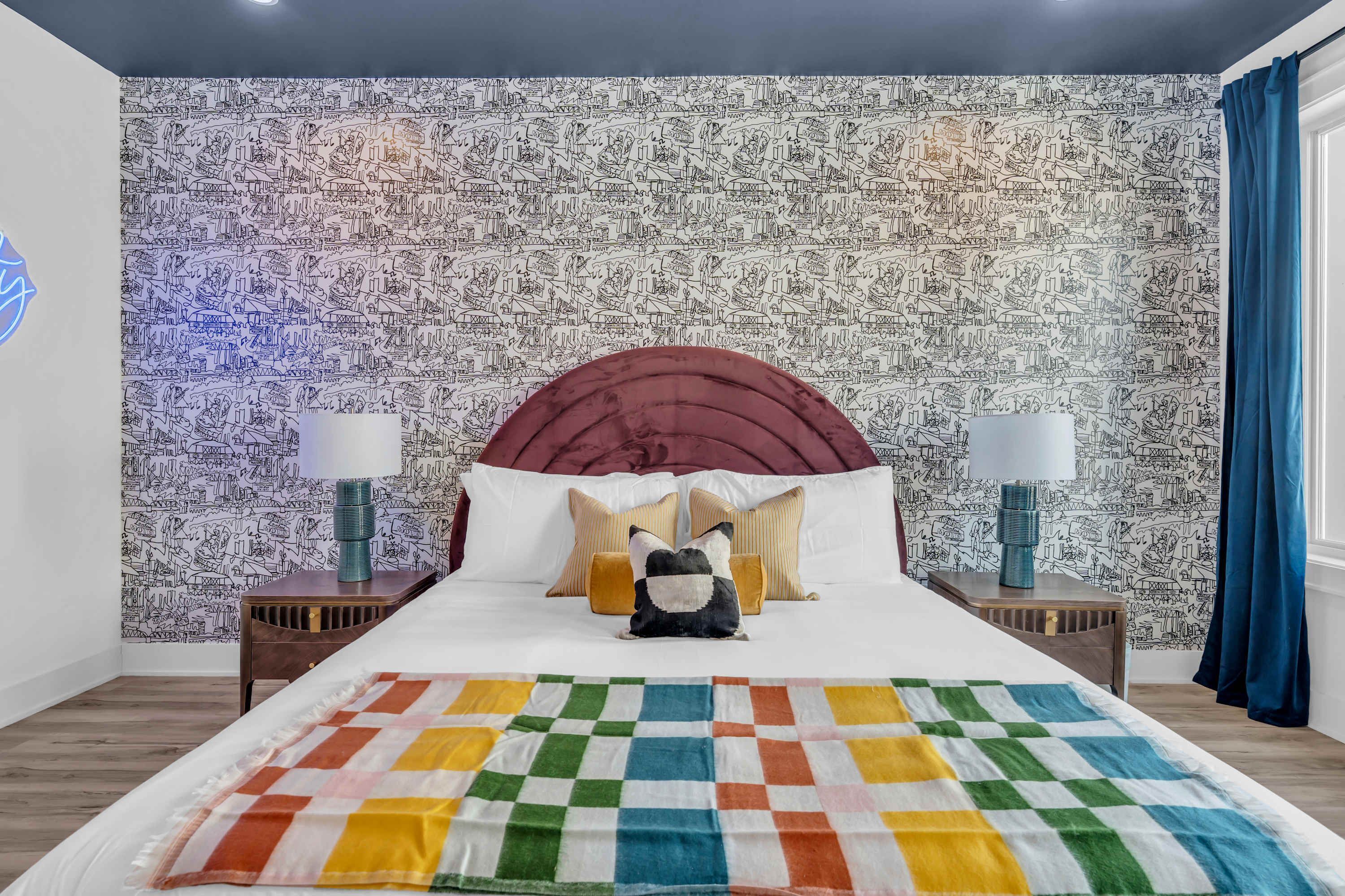 Un dormitorio con papel tapiz estampado, un cabecero rojo, dos lámparas de noche, una manta de colores y una almohada decorativa.