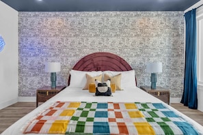 Quarto com papel de parede estampado, cabeceira vermelha, duas luminárias de apoio, manta colorida e almofada decorativa.