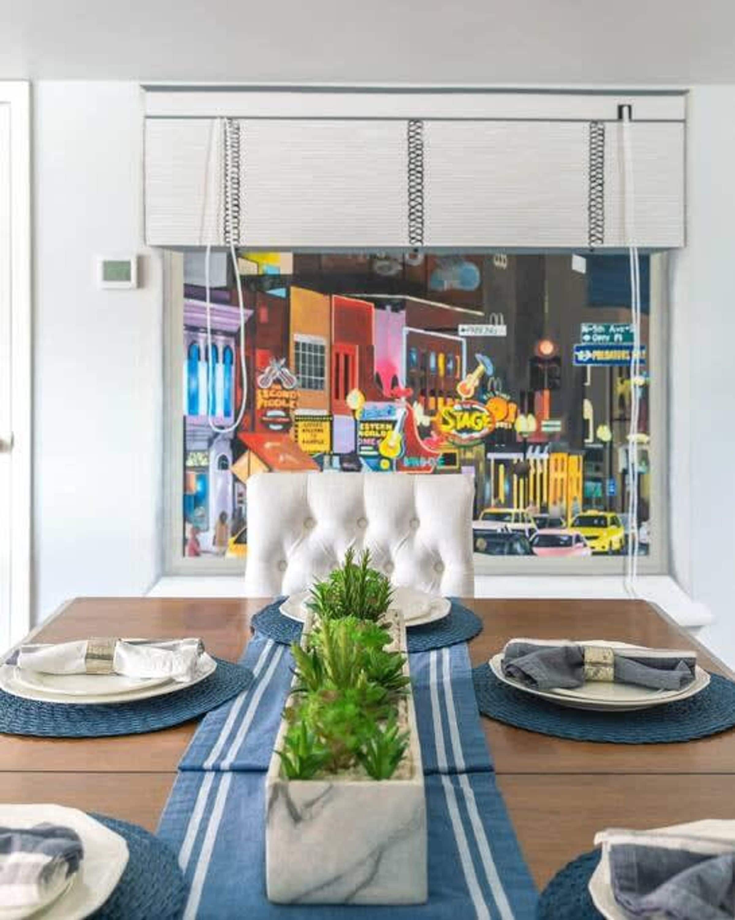 Un comedor con una mesa dispuesta para cuatro personas, una planta en maceta y vista a una calle colorida a través de una ventana.