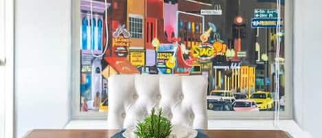 Un comedor con una mesa dispuesta para cuatro personas, una planta en maceta y vista a una calle colorida a través de una ventana.