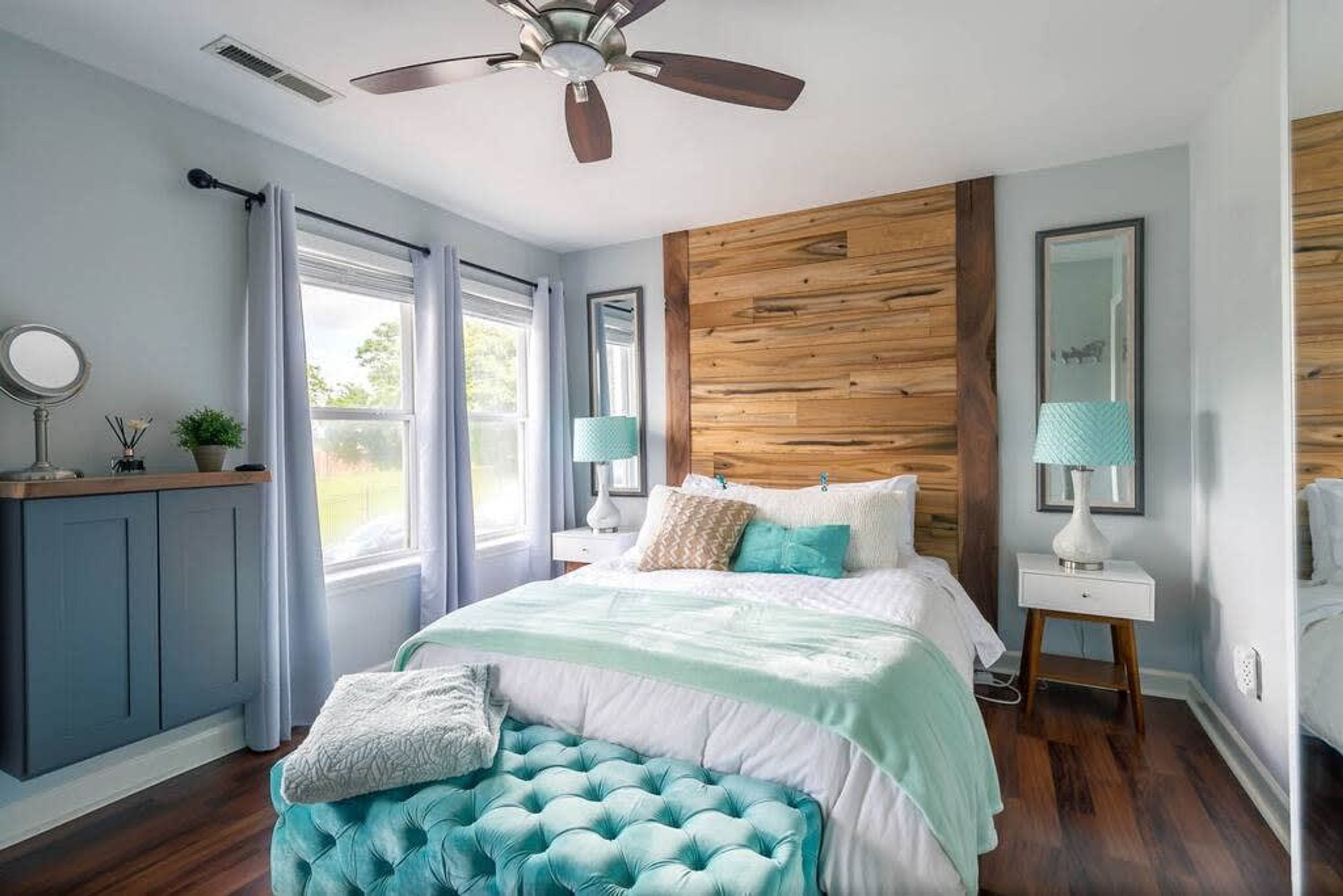 Une chambre à coucher comprenant un lit, un ventilateur de plafond, un mur d’accent en bois, une fenêtre et une table de chevet avec une lampe.