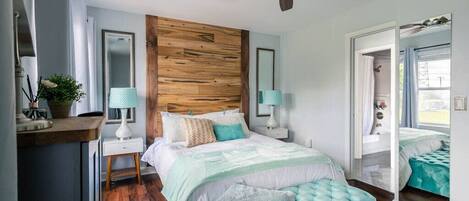 Un dormitorio con cabecera de madera, cama con reposapiés acolchado, dos mesitas de noche con lámparas, un espejo y un ventilador de techo.