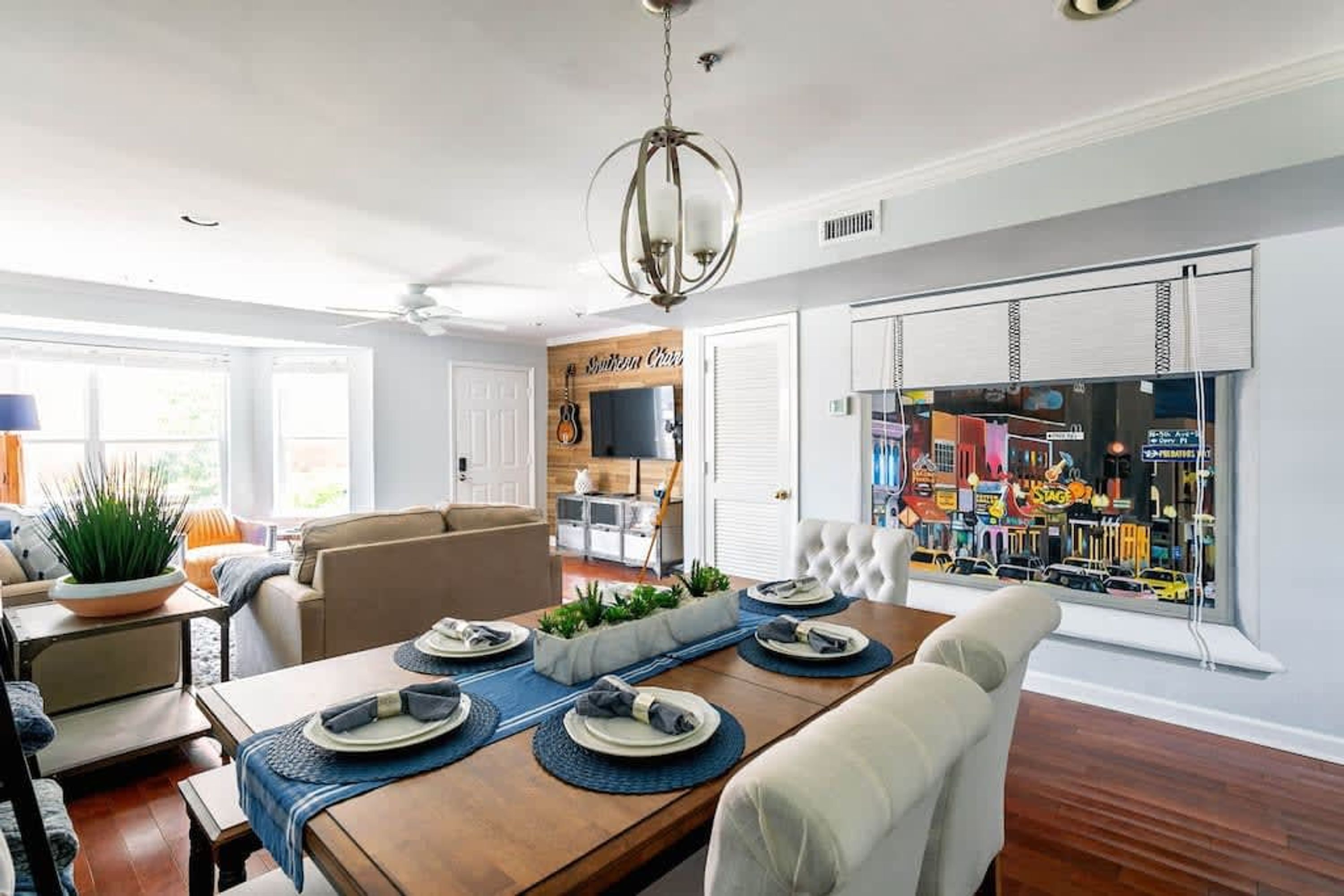 A dining area with a wooden table set for four, a ceiling fan, and a view of a street through a window with blinds.
