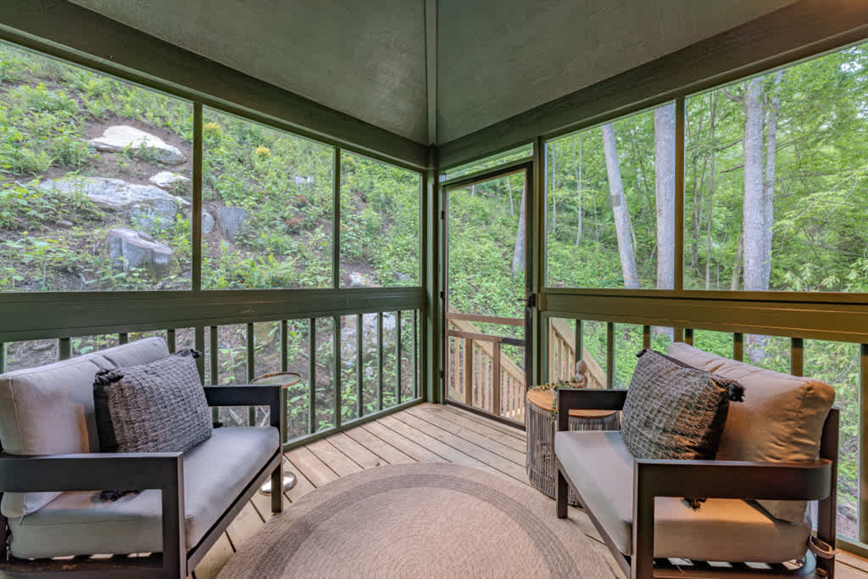 A covered outdoor seating area with two cushioned chairs and a small table, overlooking a forested landscape.