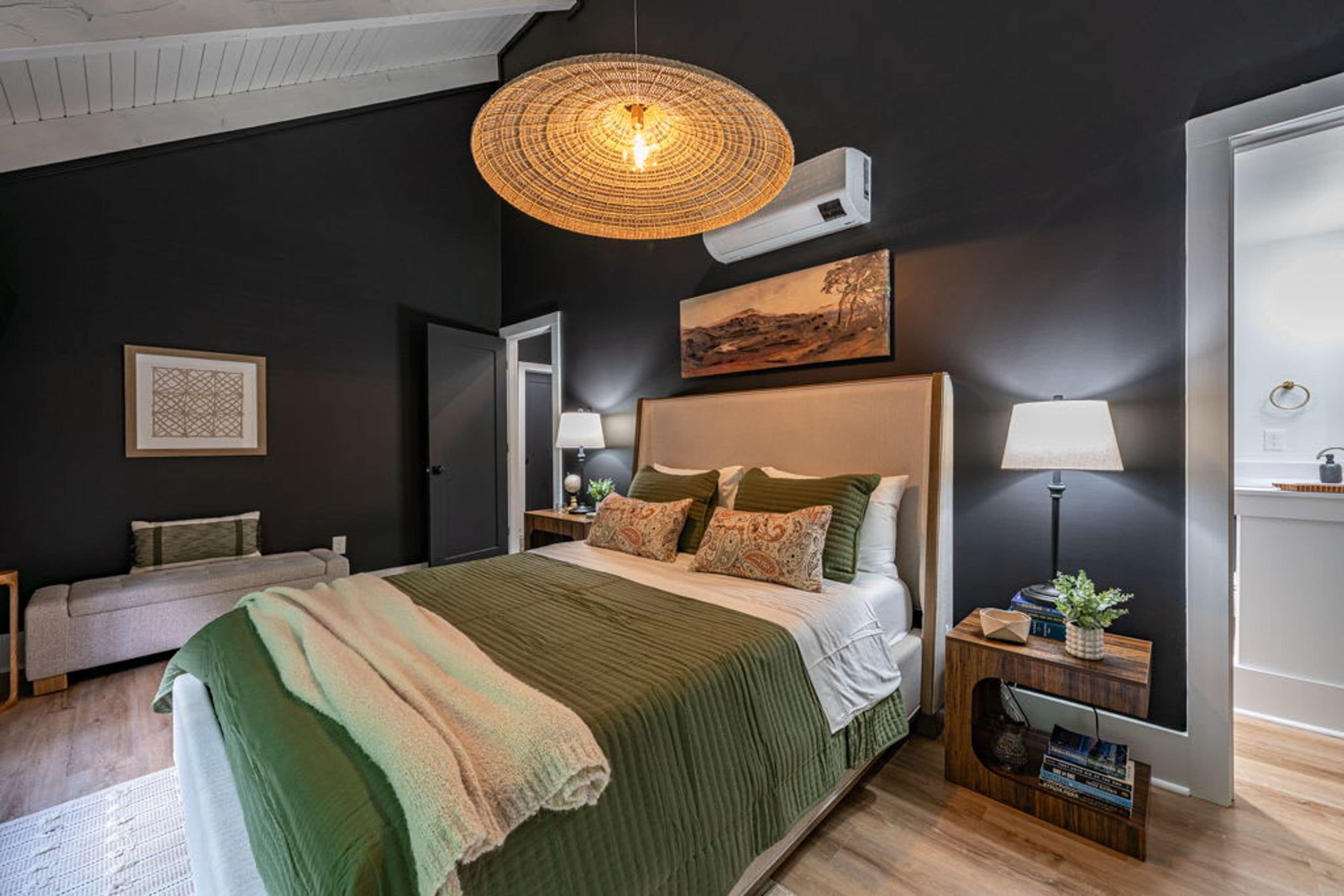 A modern bedroom with a large bed, a hanging light fixture, a painting, and a bedside table with a lamp.