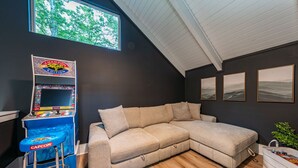 Ein Zimmer mit großem Fenster, einer Spielkonsole, einem beige farbenen Ecksofa und drei gerahmten Bildern an der Wand.