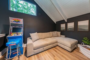 Ein Zimmer mit großem Fenster, einer Spielkonsole, einem beige farbenen Ecksofa und drei gerahmten Bildern an der Wand.