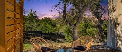 An outdoor seating area with two wicker chairs and a glass table, surrounded by trees and a wooden fence.