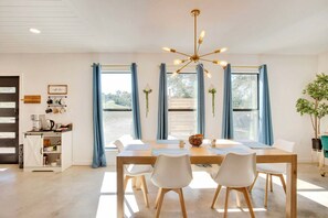 A dining area with a wooden table, white chairs, a chandelier, and blue curtains. - Hill Country Hideaway Modern Barndominium for 6 (Dripping Springs)