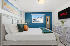 Ein Schlafzimmer mit Bett, Kissen, einem Fenster, einem Schminktisch und einem Fernseher, der 'NETFLIX' anzeigt.