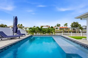 A swimming pool with lounge chairs and umbrellas, surrounded by a white fence and palm trees. - Waterfront Heated Pool Home Gym Fire Pit (Tampa)