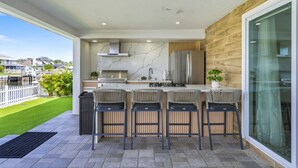 A modern outdoor kitchen with a marble backsplash, stainless steel appliances, and a large window overlooking a residential area. - Waterfront Heated Pool Home Gym Fire Pit (Tampa)