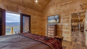 Interior de una cabaña de madera con una cama, un televisor y vistas al exterior.