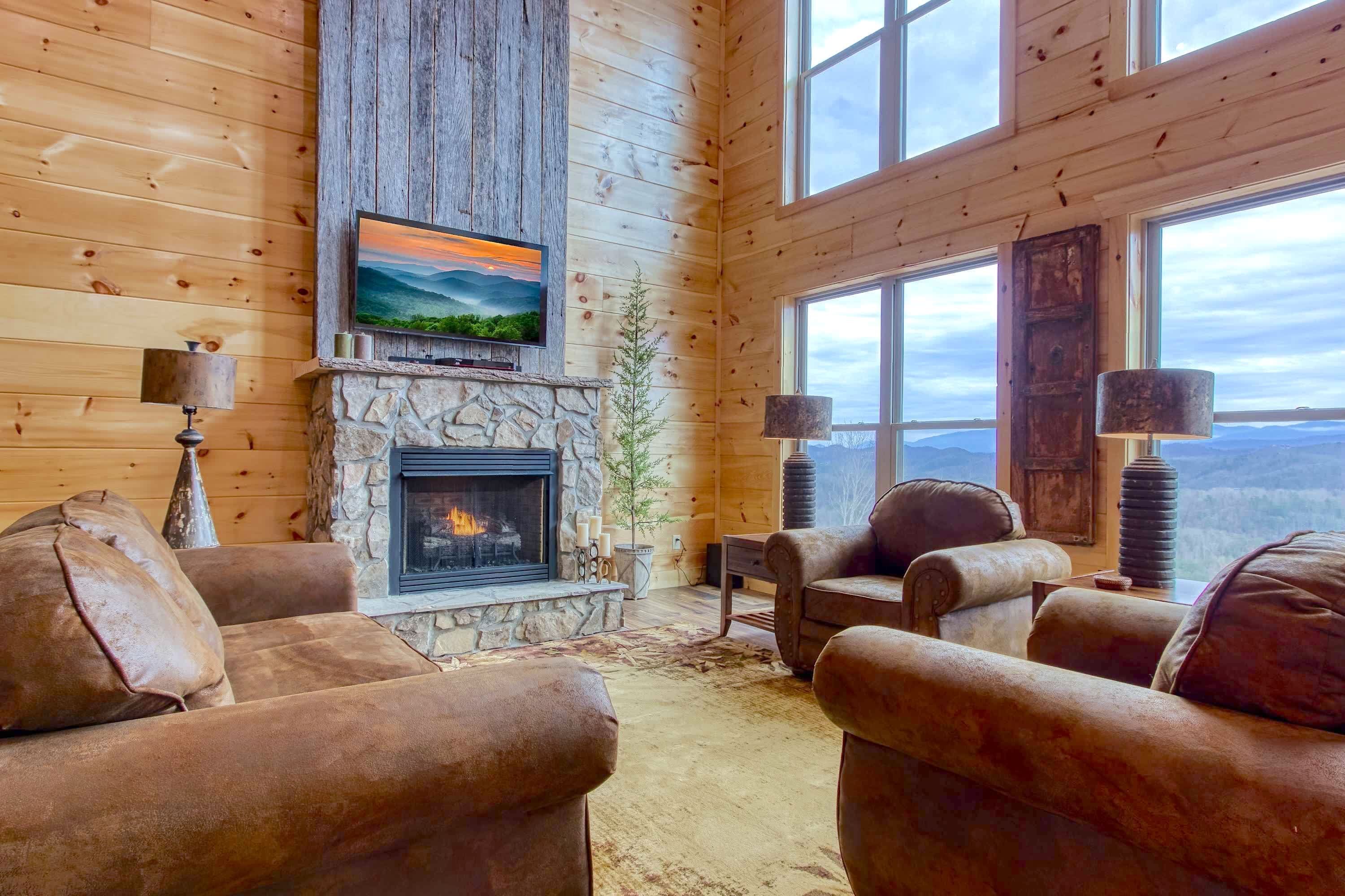 A cozy log cabin interior with a stone fireplace, leather sofas, a television mounted on a wooden wall, and a large window offering a view of the mountains.