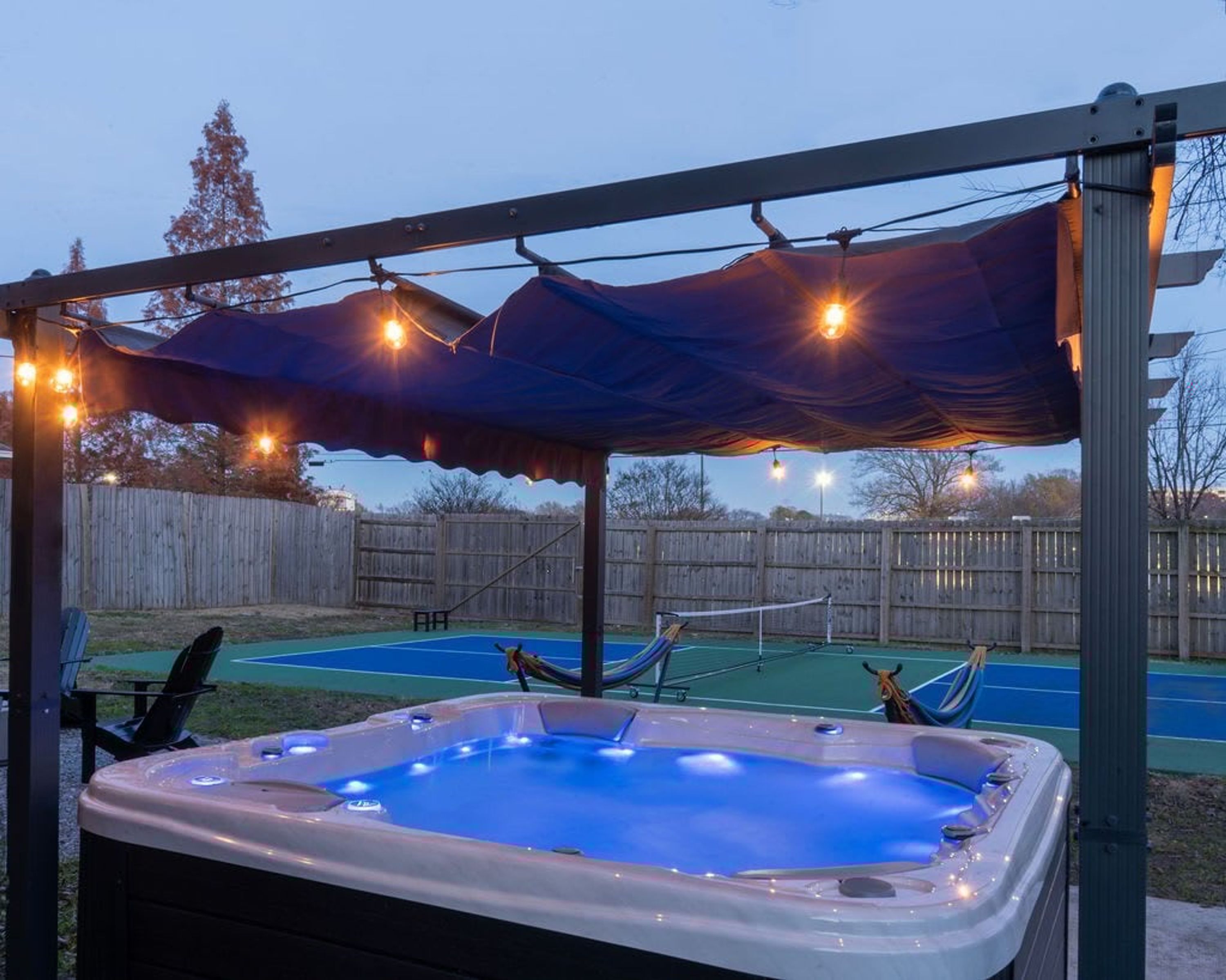 A hot tub under a covered area with string lights, a tennis court, and a hammock.