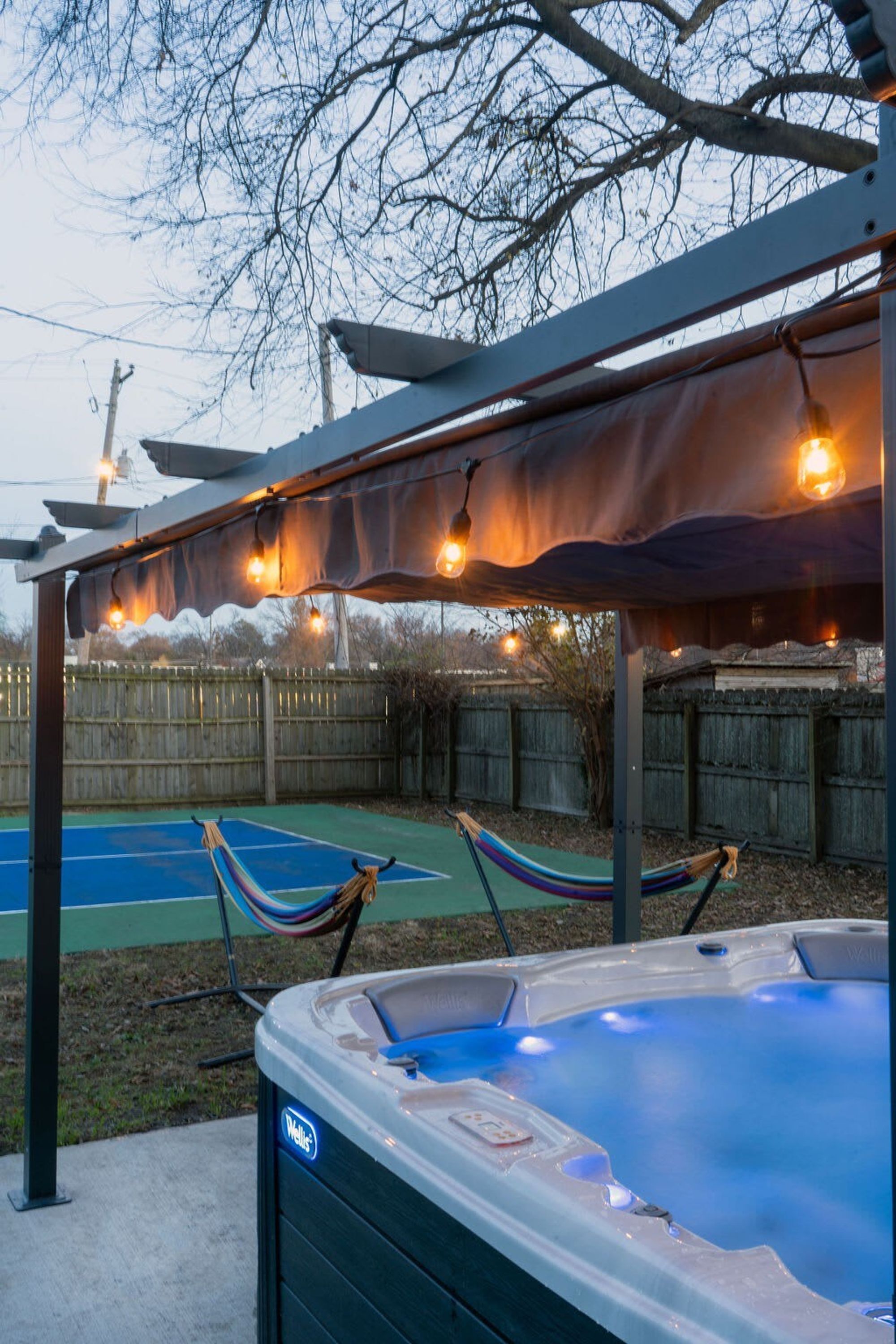A hot tub under a pergola with string lights, a hammock, and a tennis court in the background.