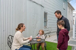 A family of three at a round metal table with a plate of food, seated outside a house with a corrugated metal wall.