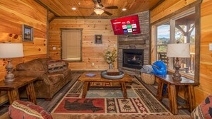 A cozy cabin interior with a wooden floor, a fireplace, a ceiling fan, and a mounted TV displaying streaming services. - Modern Cabin Movie Theater Hot Tub Game Room (Gatlinburg)