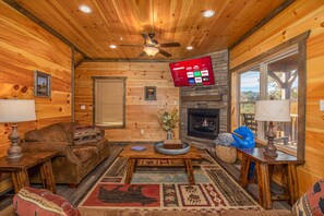 Ein gemütliches Blockhauskaminzimmer mit Holzboden, Kamin, Deckenventilator und einem an der Wand befestigten Fernseher, der Streamingdienste anzeigt.