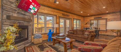 A cozy cabin interior with a stone fireplace, wooden furniture, and a large flat-screen TV mounted on the wall.