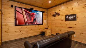 A wooden-paneled room with a large flat-screen TV mounted on the wall, a small wooden table, and a leather sofa. - Modern Cabin Movie Theater Hot Tub Game Room (Gatlinburg)