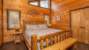 A wooden cabin bedroom with a bed, a bench, a nightstand, a window, and a door. - Modern Cabin Movie Theater Hot Tub Game Room (Gatlinburg)