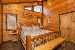 Ein Schlafzimmer in einer Holzhütte mit einem Bett, einer Bank, einem Nachttisch, einem Fenster und einer Tür.
