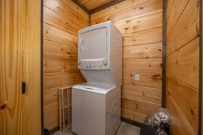 A compact laundry room with a stacked washer and dryer, wooden walls, and a small vent. - Modern Cabin Movie Theater Hot Tub Game Room (Gatlinburg)