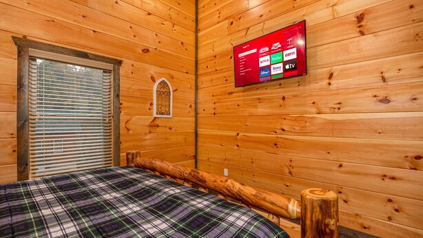 A wooden cabin room with a bed, a window with blinds, and a mounted TV displaying streaming services. - Modern Cabin Movie Theater Hot Tub Game Room (Gatlinburg)