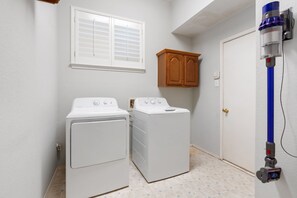 A laundry room with two white washing machines, a wooden cabinet, a window with blinds, and a vacuum cleaner mounted on the wall. - Heated Pool Hot Tub Sleeps 16 Putting Green (McKinney)