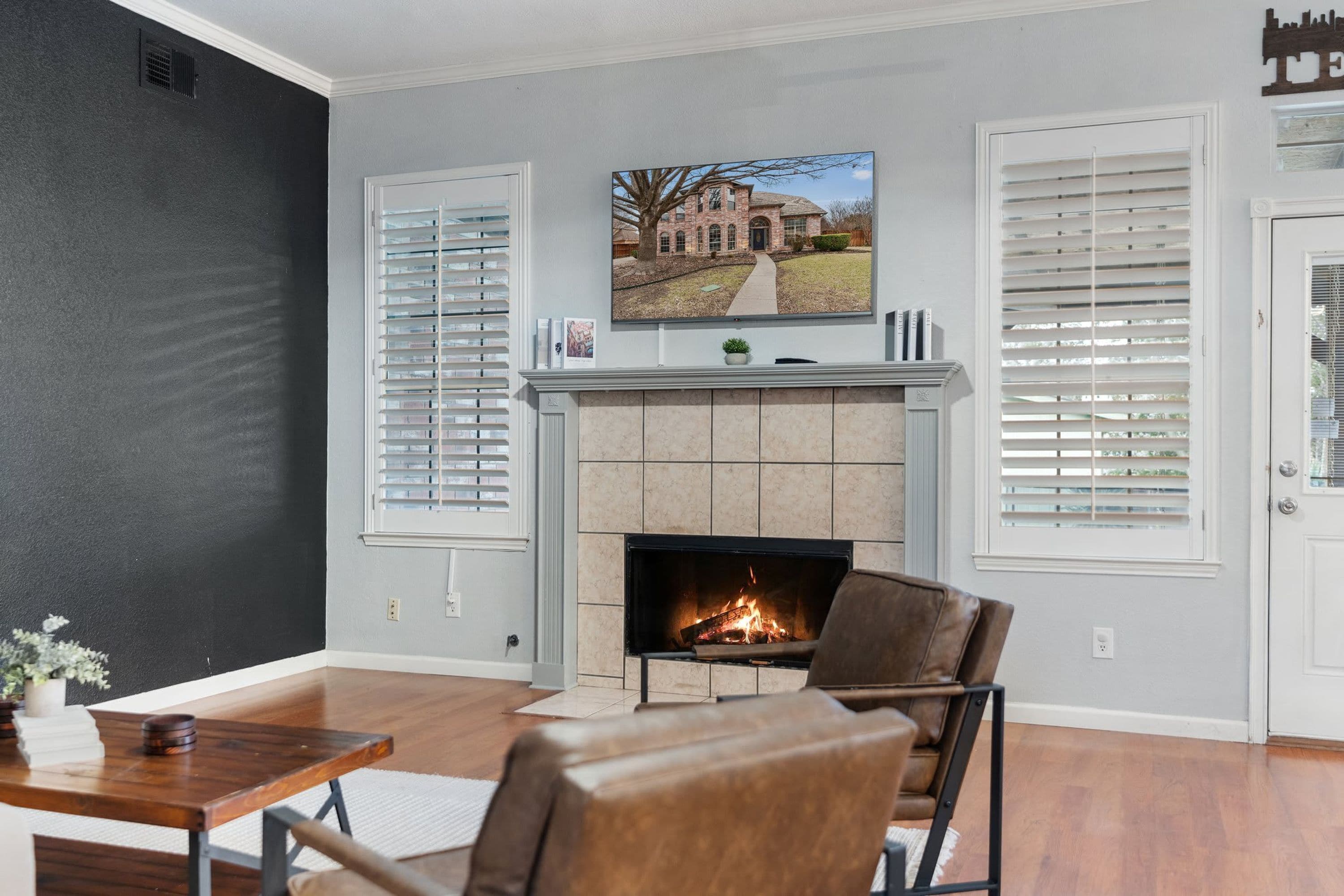 A living room with a fireplace, two windows with blinds, a television mounted above the fireplace, and a door with a decorative sign.
