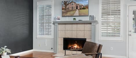 A living room with a fireplace, two windows with blinds, a television mounted above the fireplace, and a door with a decorative sign.