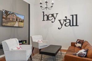 A living room with two white armchairs, a brown sofa, a black coffee table, and a wall-mounted TV displaying a house. - Heated Pool Hot Tub Sleeps 16 Putting Green (McKinney)