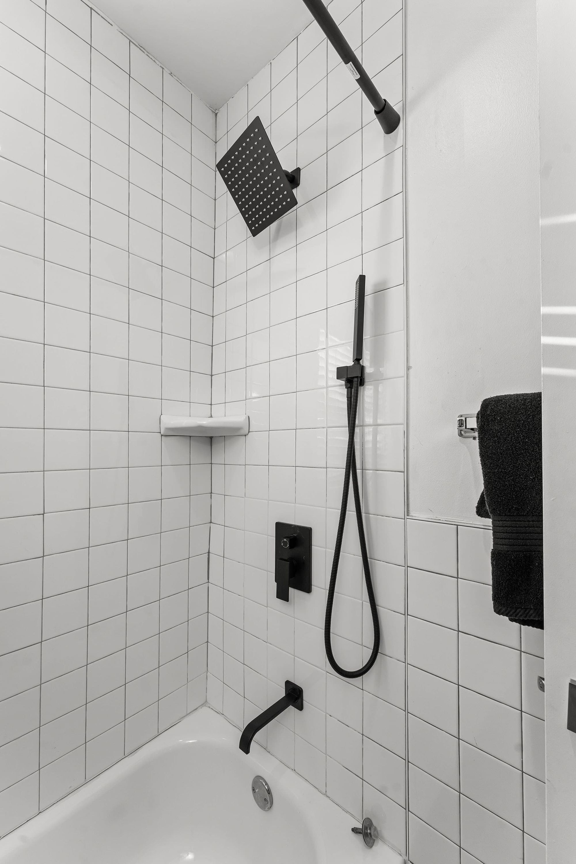 Ein Badezimmer mit weiß gefliesten Wänden, einem schwarzen Duschkopf, einer Handbrause und einem schwarzen Handtuch, das an der Wand hängt.