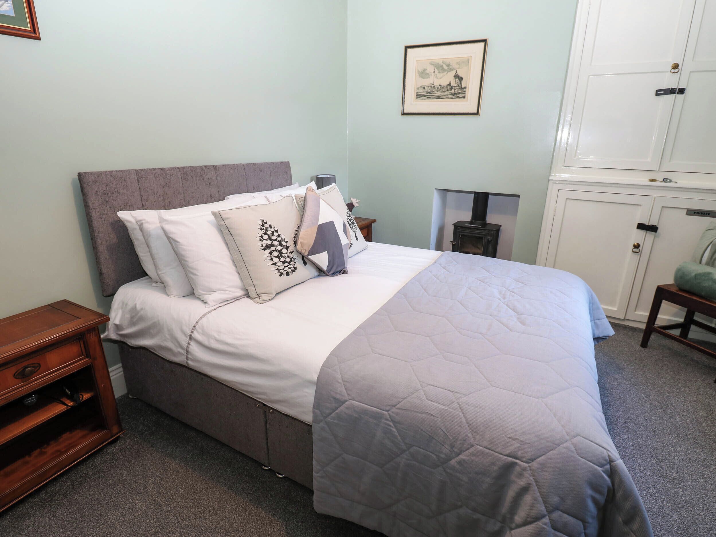 Un lit bien fait, avec une couverture matelassée, une tête de lit et des coussins décoratifs. Un tableau encadré est accroché au mur au-dessus du lit, et un meuble avec une étiquette indiquant « PRIVÉ » se trouve à droite.