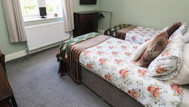 A bedroom with two beds, a radiator, a window with curtains, a flat-screen TV, and a wooden dresser. - Prudhoe Cottage (Prudhoe)