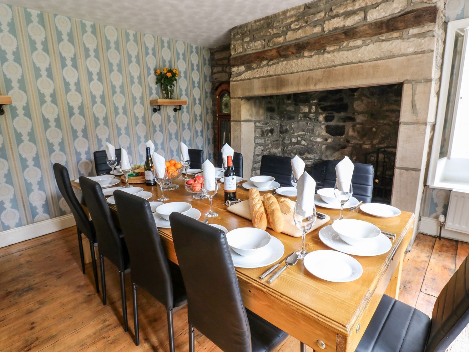 A dining room with a long wooden table set for a meal, black chairs, a stone fireplace, and patterned wallpaper.