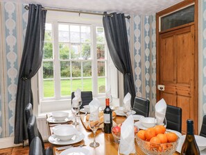A dining room with a table set for two, a window with curtains, and a view of greenery outside. - Prudhoe Cottage (Prudhoe)