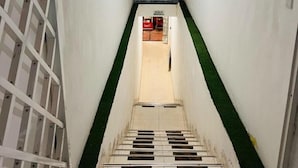 Un pasillo estrecho con suelo de baldosas, una sección alfombrada en verde y una pared de ladrillo al final.