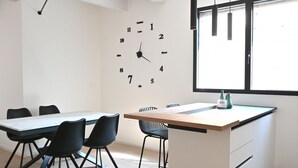 Eine moderne Küche mit einer großen Wanduhr, einem Esstisch mit Stühlen und einem Fenster mit Jalousien.