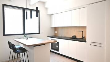 A modern kitchen with white cabinets, a black countertop, and a built-in oven. There is a small island with a bar stool and a window above the sink.