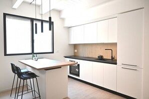 A modern kitchen with white cabinets, a black countertop, and a built-in oven. There is a small island with a bar stool and a window above the sink. - SE310 – Senigallia, stylish apartment with air conditioning, private garage and outdoor courtyard (Senigallia)