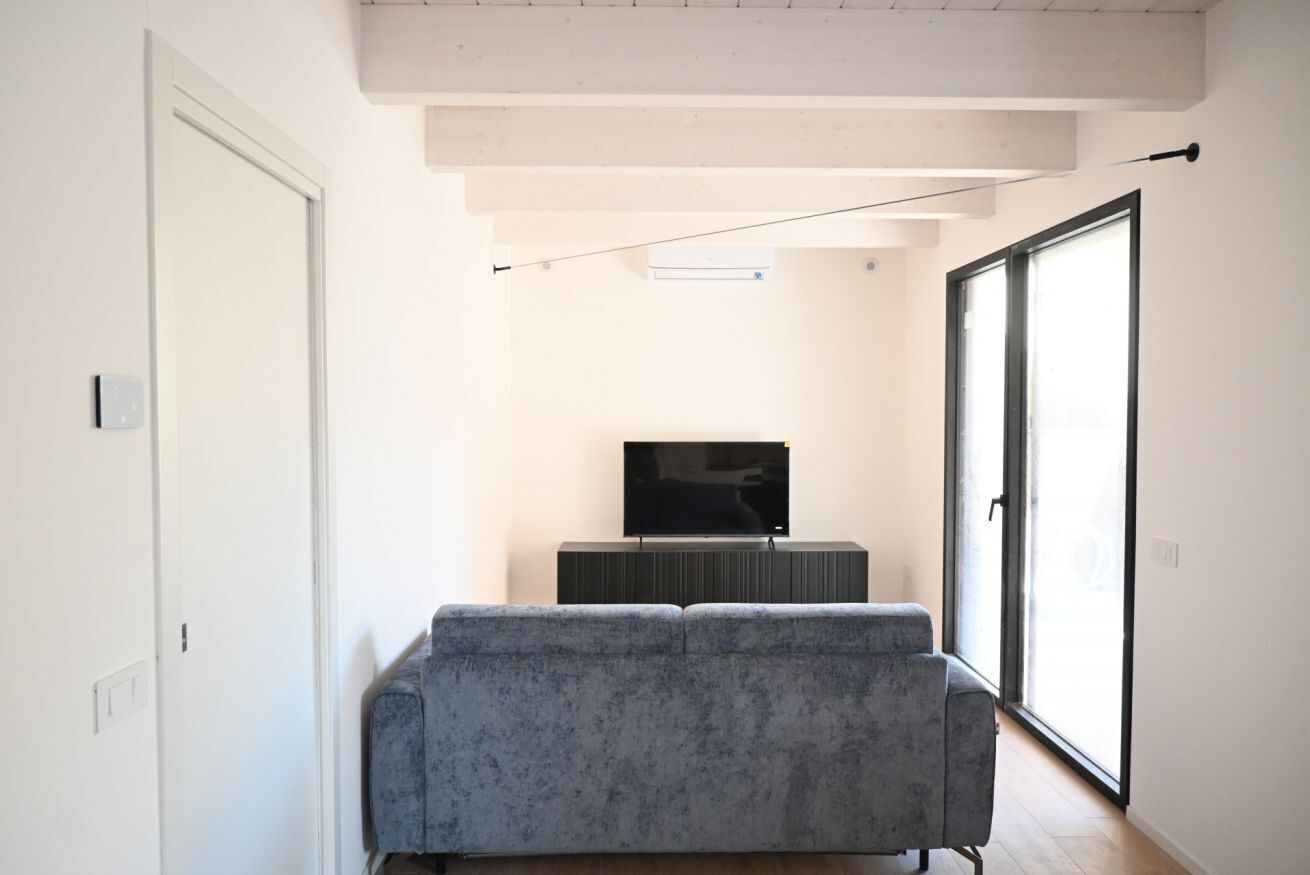 A modern living room with a grey sofa, a flat-screen TV on a black stand, and a large sliding glass door leading to a balcony.