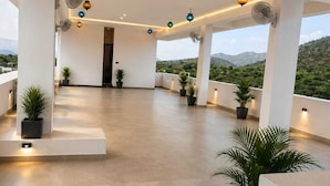 A spacious indoor area with ceiling fans, potted plants, and a view of the outdoors. - The Art of Field (Udaipur)