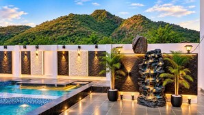 A swimming pool with a waterfall feature, surrounded by stone walls and potted plants, with a view of a mountainous landscape. - The Art of Field (Udaipur)